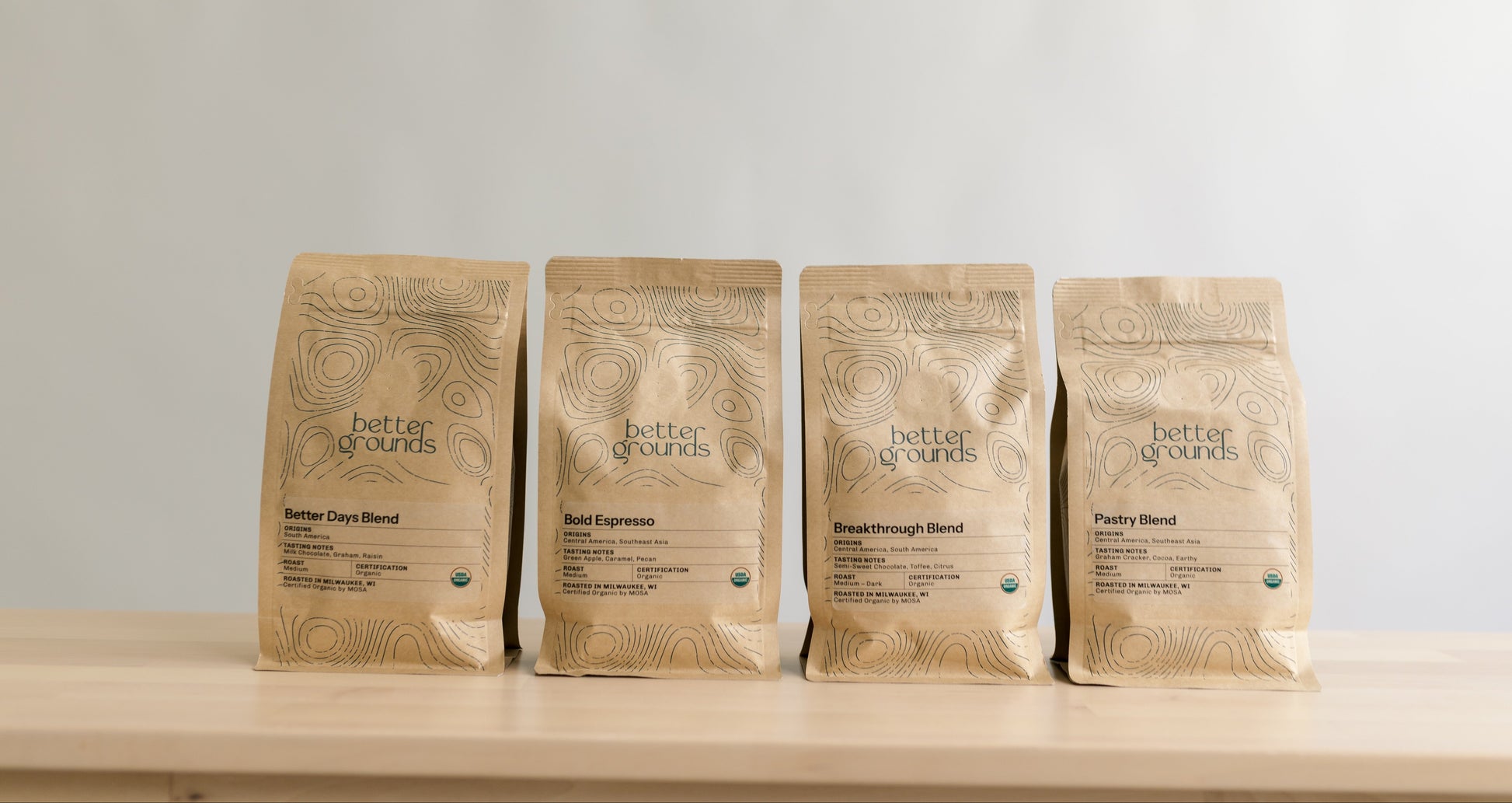 Four packages of 'Better Grounds' coffee on a wooden surface with a light gray background